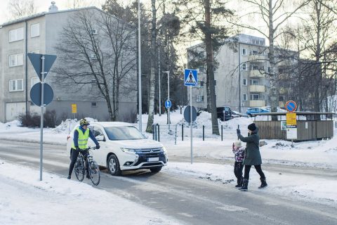 Autoilija ja pyöräilijä antavat tietä suojatiellä käveleville jalankulkijoille. Kävelijät tervehtivät autoa ja pyöräilijää kulkiessaan tien yli.