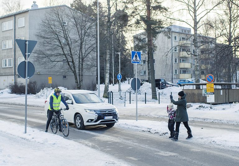 Autoilija ja pyöräilijä antavat tietä suojatiellä käveleville jalankulkijoille. Kävelijät tervehtivät autoa ja pyöräilijää kulkiessaan tien yli.