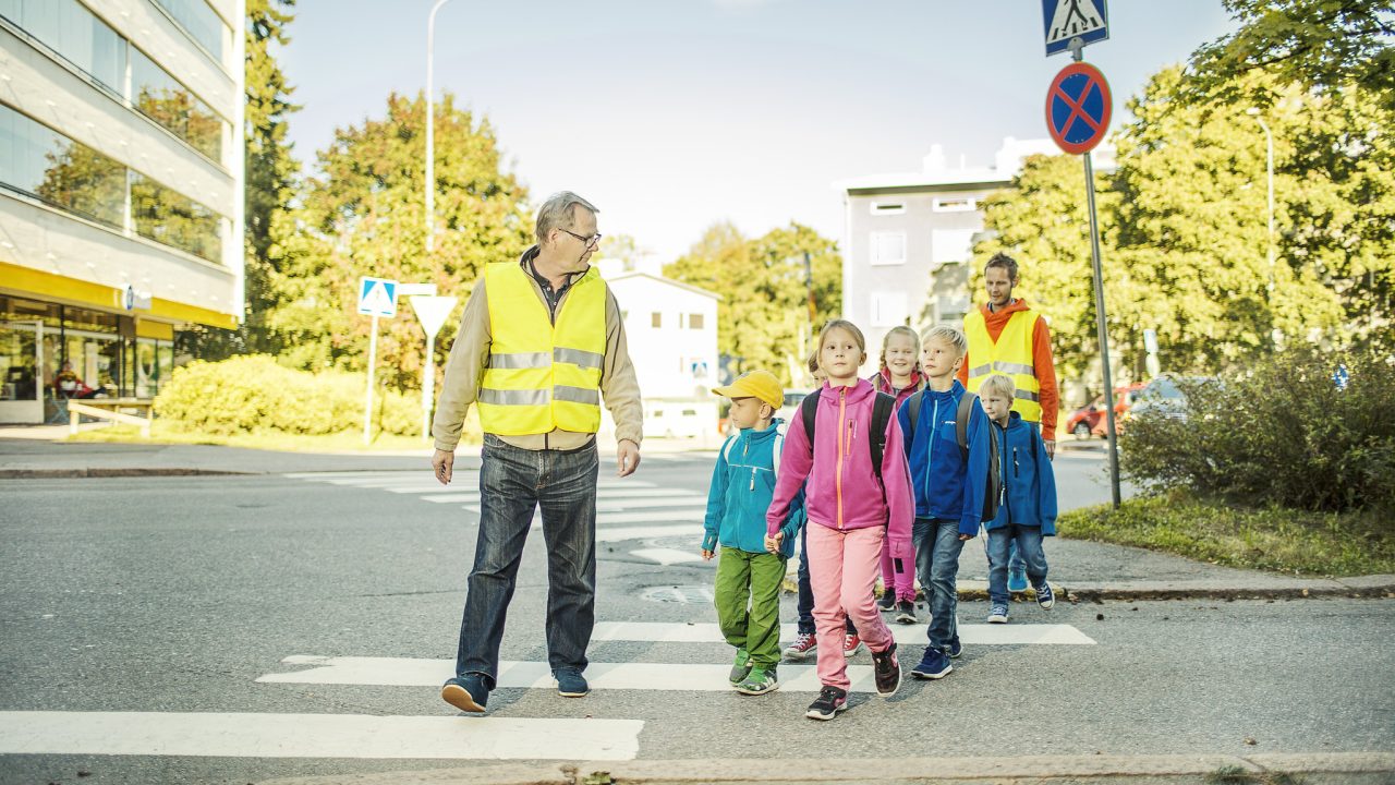 kaksi aikuista ja useita lapsia ylittämässä suojatietä yhdessä.