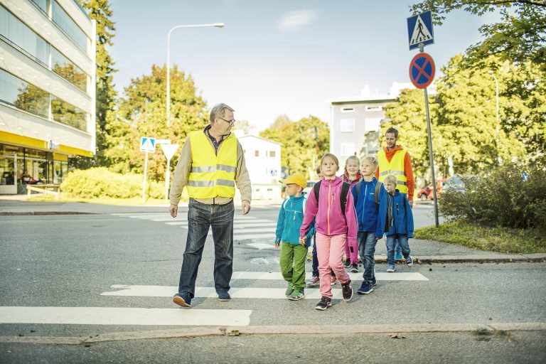 Kaksi aikuista ja useita lapsia ylittämässä suojatietä yhdessä. Aikuisilla on huomioliivit.