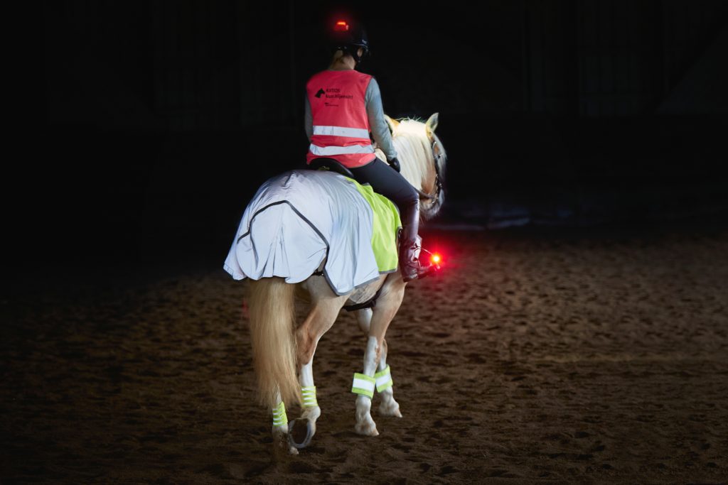 Kuvassa ratsukko, jolla ratsastajalla kypärässä punainen valo osoittamassa taaksepäin sekä myö punainen valo kiinni jalustimessa osoittamassa taaksepäin.