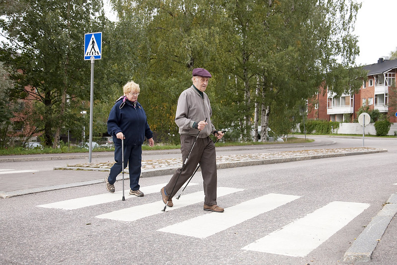 Vanhempi pariskunta ylittää suojatien. Apuna kävelysauvat ja kävelykeppi.