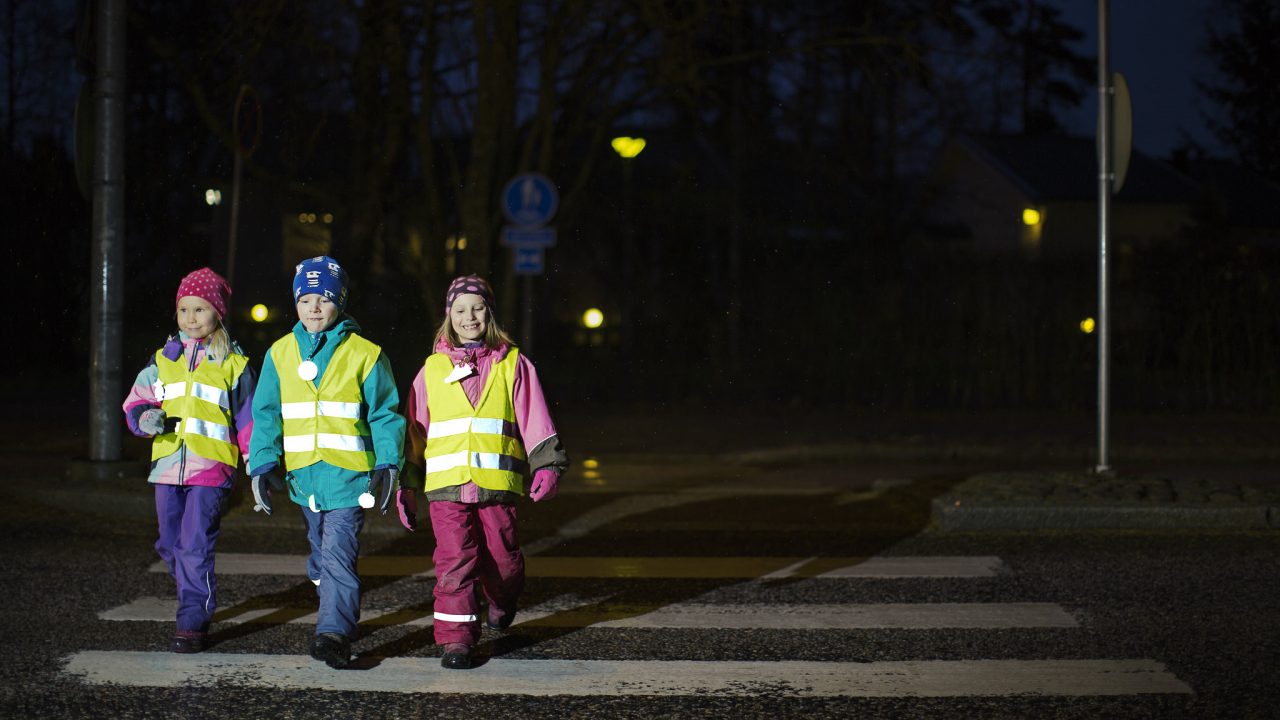 Kolme lasta ylittää suojatietä varusteinaan heijastinliivi ja heijastimia.