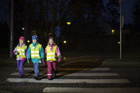 Kolme lasta ylittää suojatietä varusteinaan heijastinliivi ja heijastimia.