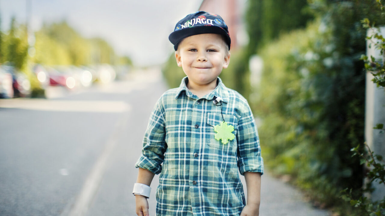 Lapsi seisoo jalkakäytävällä. Paidassa on kiinni heijastin.