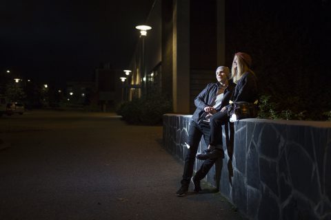 Kaksi nuorta, joista toinen istuu kiviaidan päällä ja toinen nojaamassa aitaan. Molemmilla heijastimet yllään.