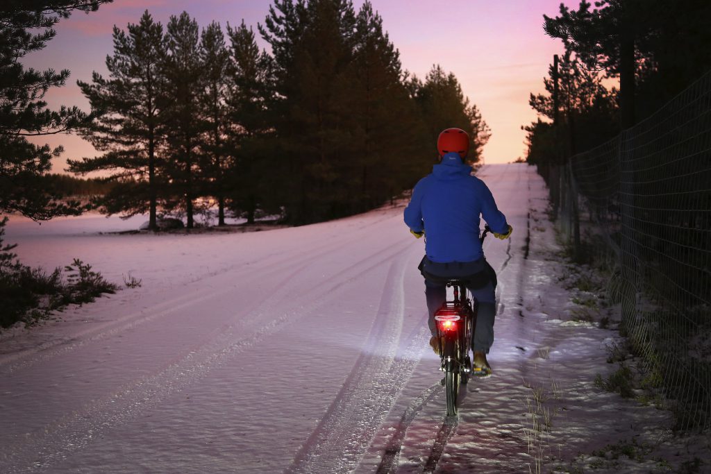 Pyöräilijä pyöräilee hämärässä, tiellä lunta. Punainen takavalo loistaa.
