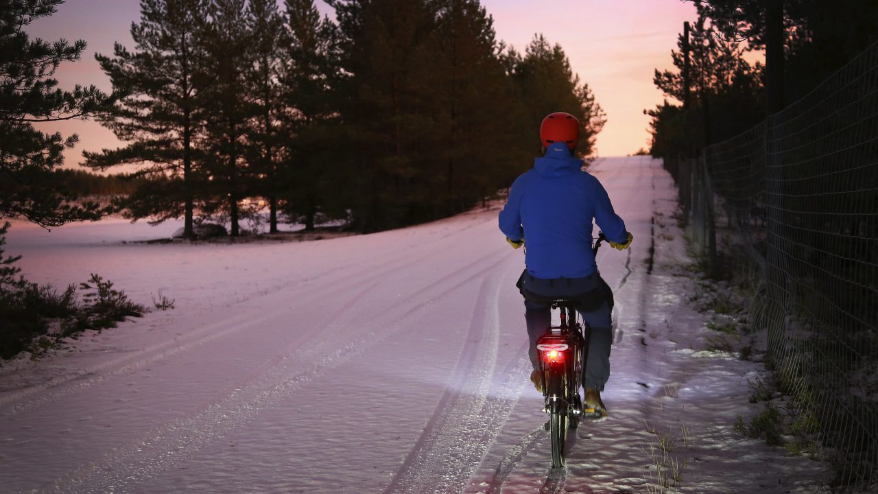 Pyöräilijä pyöräilee hämärässä, tiellä lunta. Punainen takavalo loistaa.