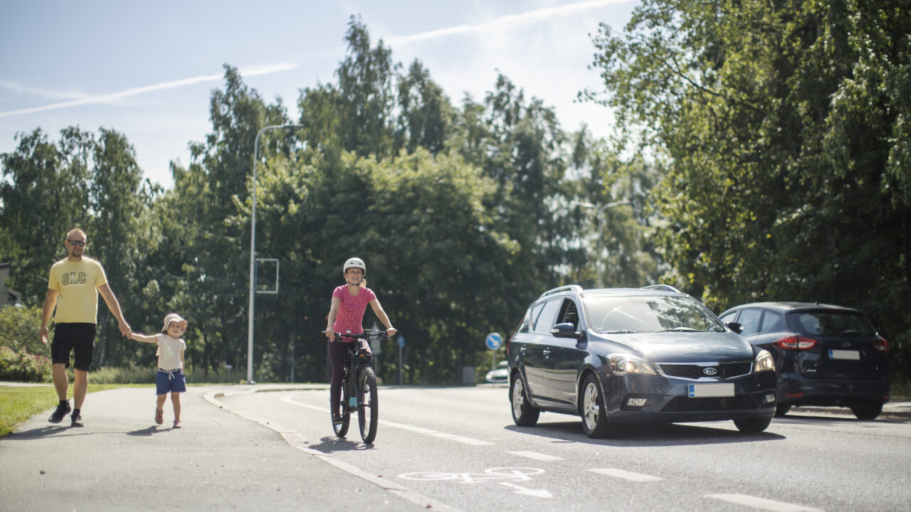 Suojatietä ylittämässä aikuinen ja lapsi. Auto ja pyöräilijä pysähtyneinä väistämistä osoittavan liikennemerkin takana.