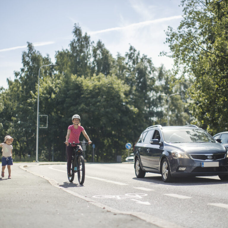 Suojatietä ylittämässä aikuinen ja lapsi. Auto ja pyöräilijä pysähtyneinä väistämistä osoittavan liikennemerkin takana.