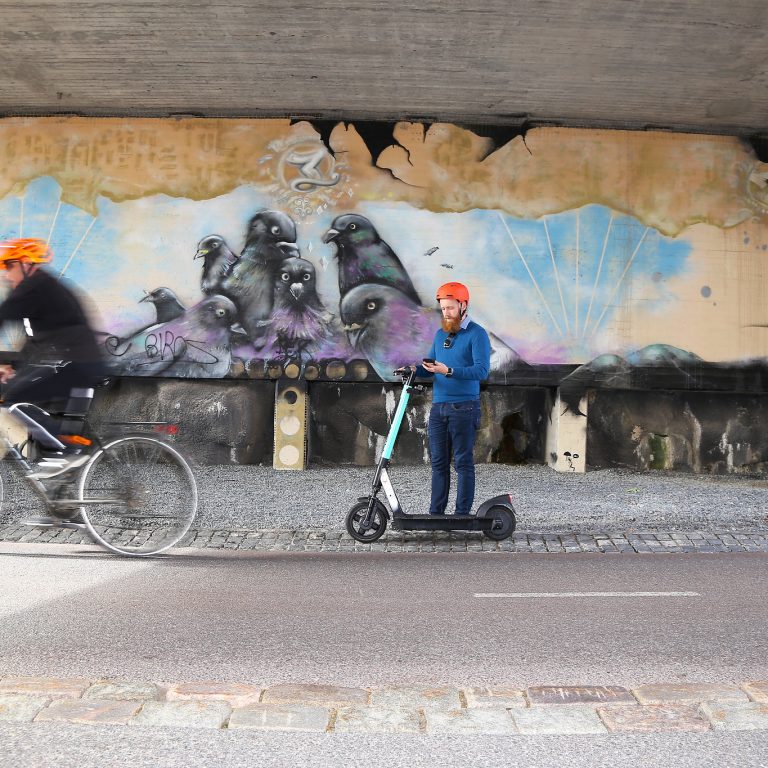 Sähköpotkulautailija on pysähtynyt käyttämään kännykkää tunnelissa tien sivuun. Pyöräilijä pyöräilee ohi.
