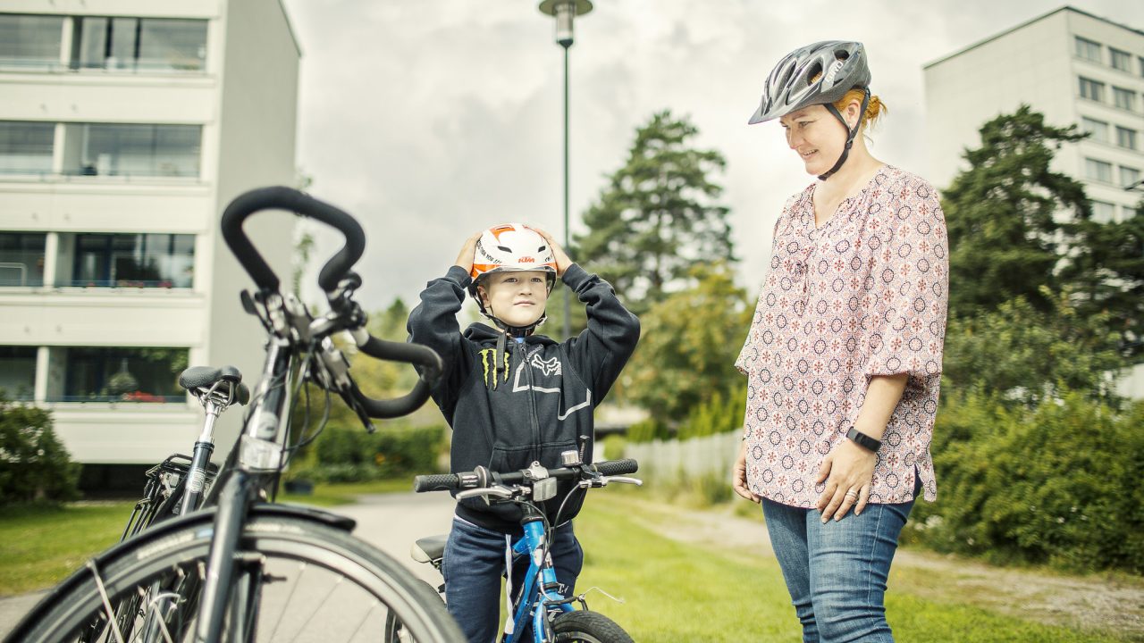 Lapsi asettaa pyöräilykypärää päähänsä ja aikuinen seuraa vieressä.