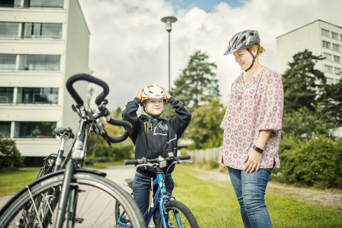 Lapsi asettaa pyöräilykypärää päähänsä ja aikuinen seuraa vieressä.