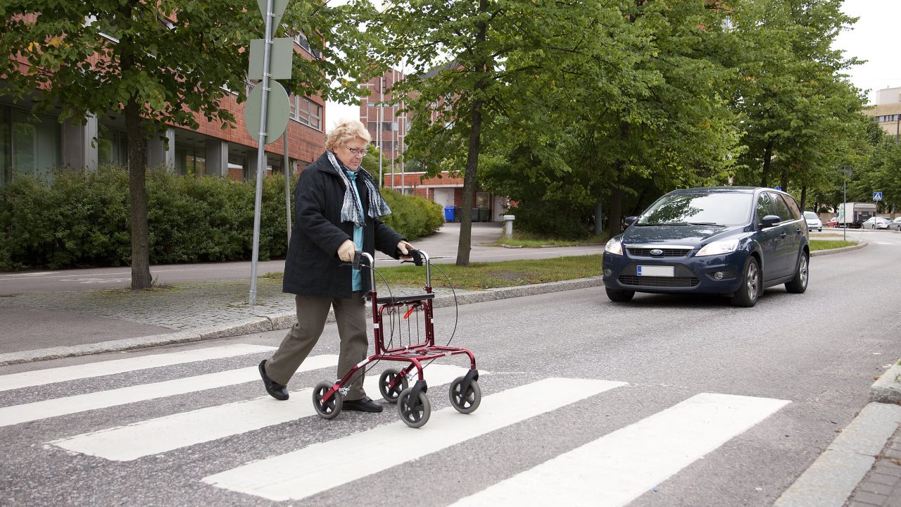 Rollaattoria käyttävä jalankulkija ylittää tietä suojatietä pitkin, autoilija antaa tietä.