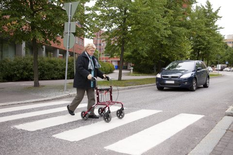 Rollaattoria käyttävä jalankulkija ylittää tietä suojatietä pitkin, autoilija antaa tietä.