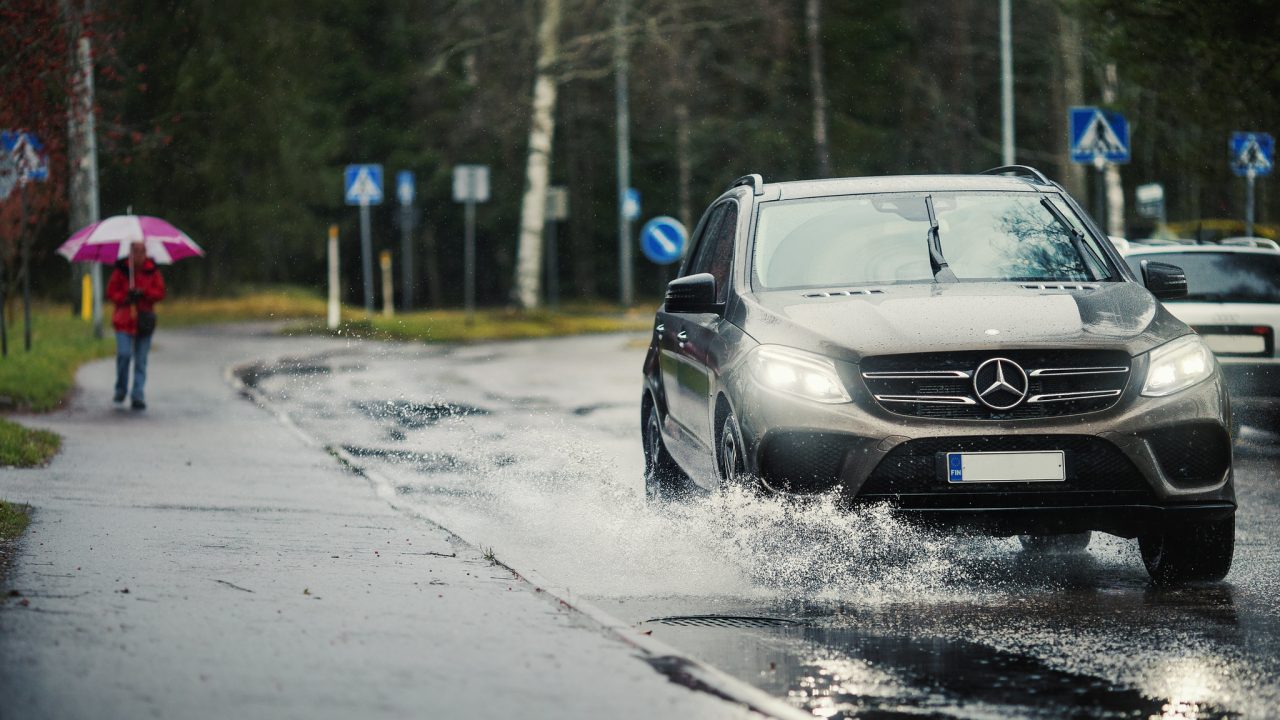 Auto ajaa sateisessa kelissä. Taustalla kävelijä sateenvarjon kanssa.