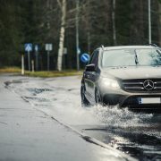 Auto ajaa sateisessa kelissä. Taustalla kävelijä sateenvarjon kanssa.