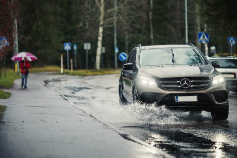 Auto ajaa sateisessa kelissä. Taustalla kävelijä sateenvarjon kanssa.