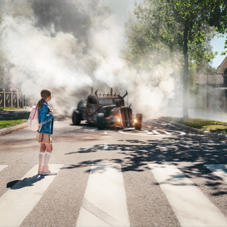 Tyttö kävelee suojatiellä. Savuttava hirviöauto lähestyy suojatietä.