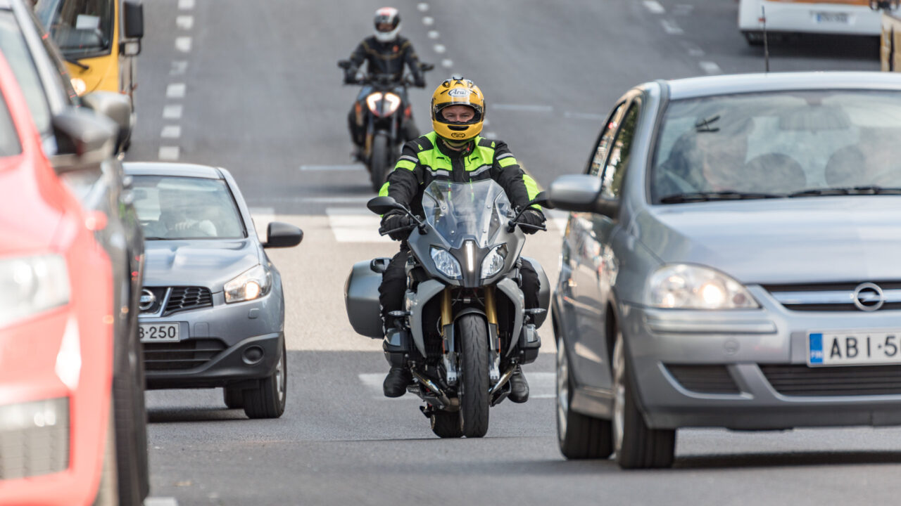 Moottoripyöräilijä matkustajineen vilkkaassa liikenteessä.
