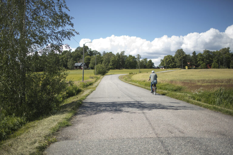 Pyöräilijä ajaa oikeaa maantien laitaa maaseutumaisessa ympäristössä.